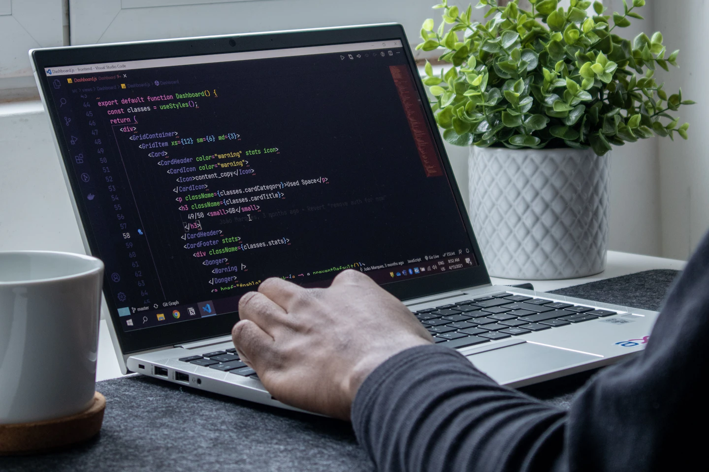 A developer working on a laptop beside a mug and a plant.