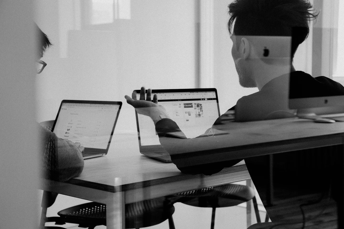 A person working across two laptops, photographed through an office window.